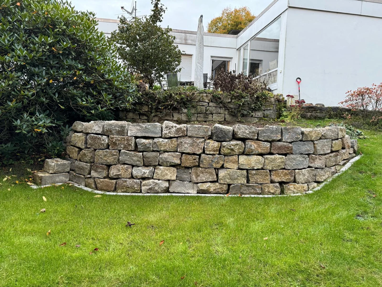 Geschwungene Natursteinmauer mit Raseneinfassung vor einem bepflanzten Hang